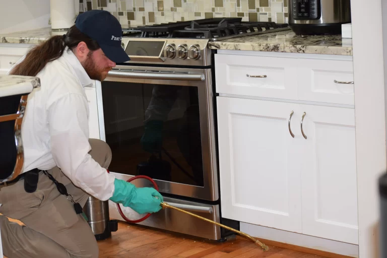 Pest control technician spraying insecticide with a long nozzle under a kitchen cabinet next to an oven in Rose Hill.